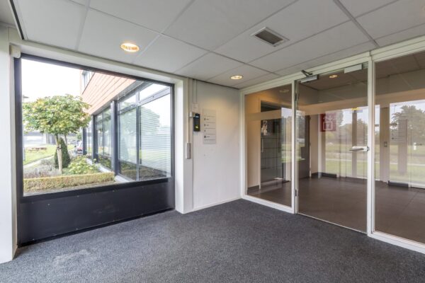 Ingang van een modern kantoorpand aan de Lage Biezenweg met glazen deuren en uitzicht op de tuin.